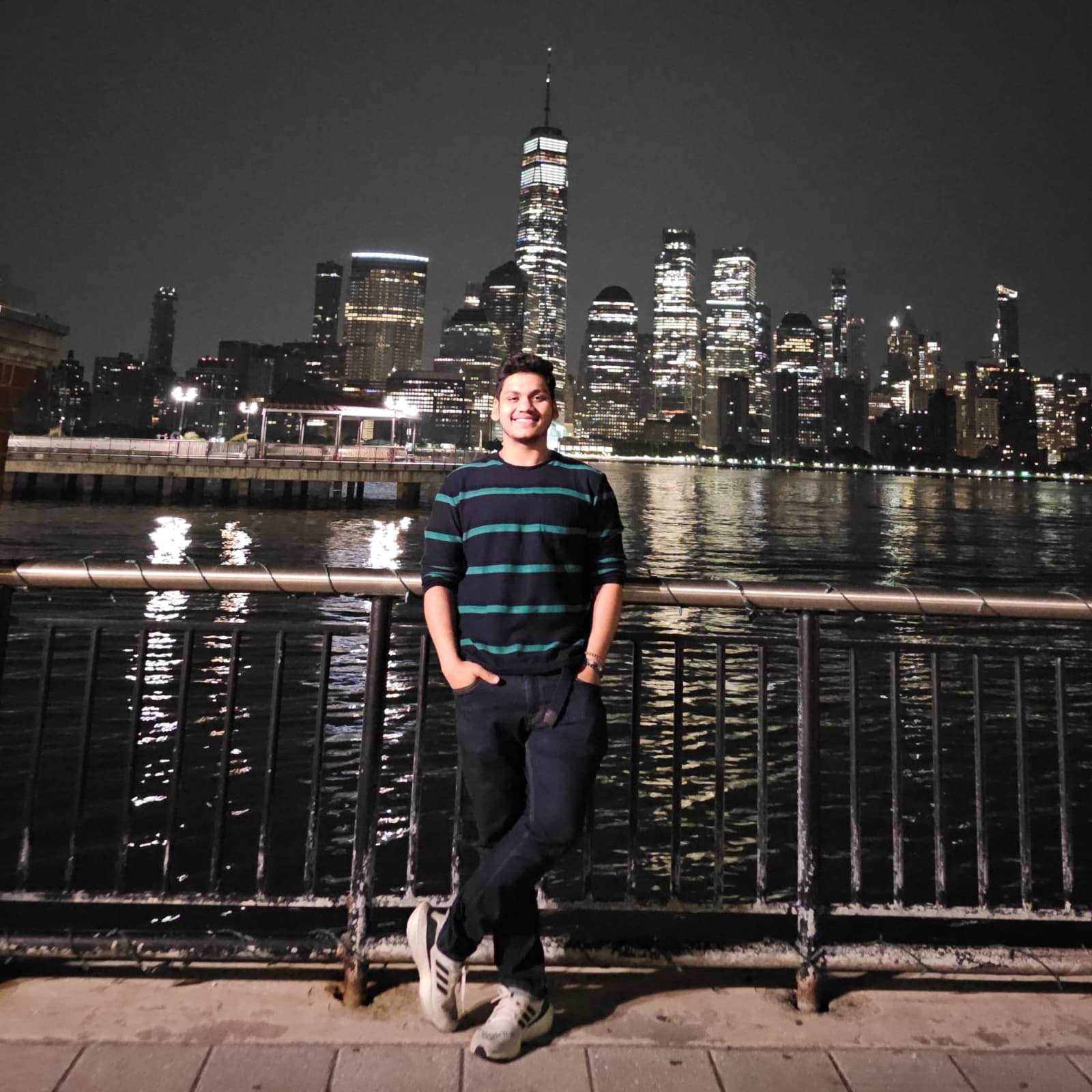 Kanishk on the Hudson waterfront at night, the Lower Manhattan skyline lit up behind him